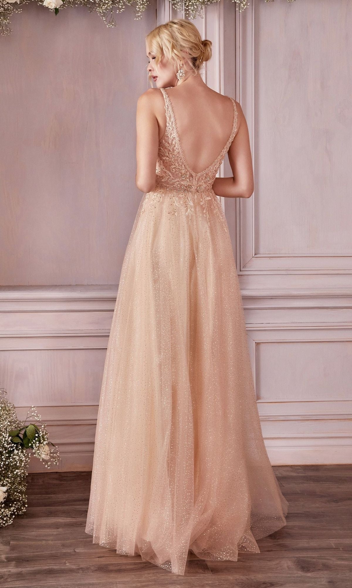 A woman faces away in a champagne Beaded-Bodice Long Pastel A-Line Prom Dress CD0196, a sparkling floor-length gown with a sleeveless design and deep V-back, standing in a room adorned with floral accents.